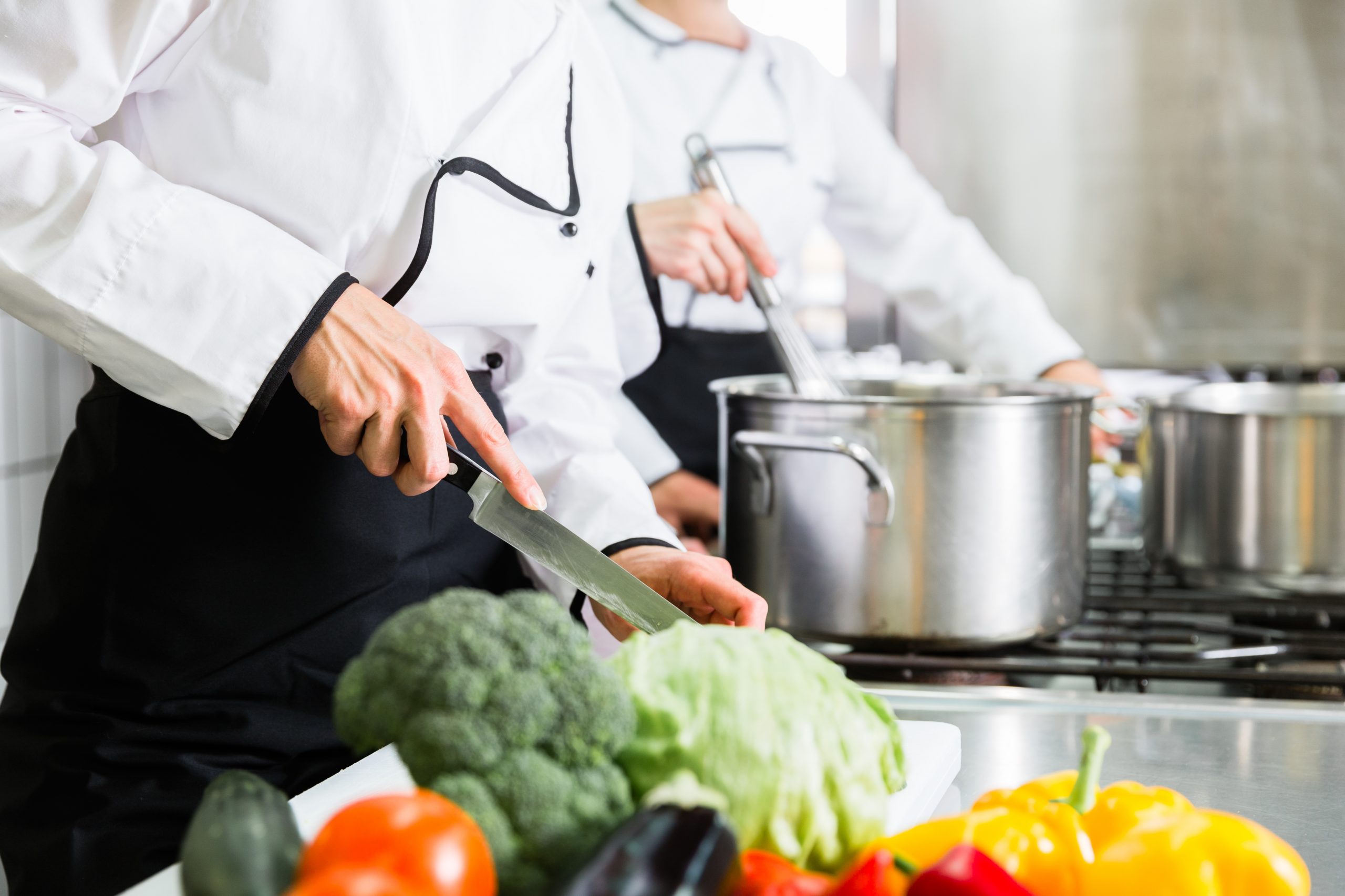 Zwei Köche stehen in einer Großküche. Eine Person rührt in einem Topf, während im Vordergrund frisches Gemüse geschnitten wird.